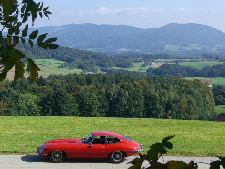 Roter Jaguar E-Type auf einer Landstraße, im Hintergrund bewaldete Hügel