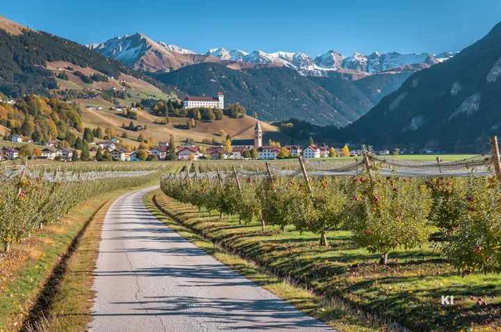KI-Symbolbild: Apfelgärten im Vinschgau mit Dorf, Kloster am Hang und verschneiten Gipfeln
