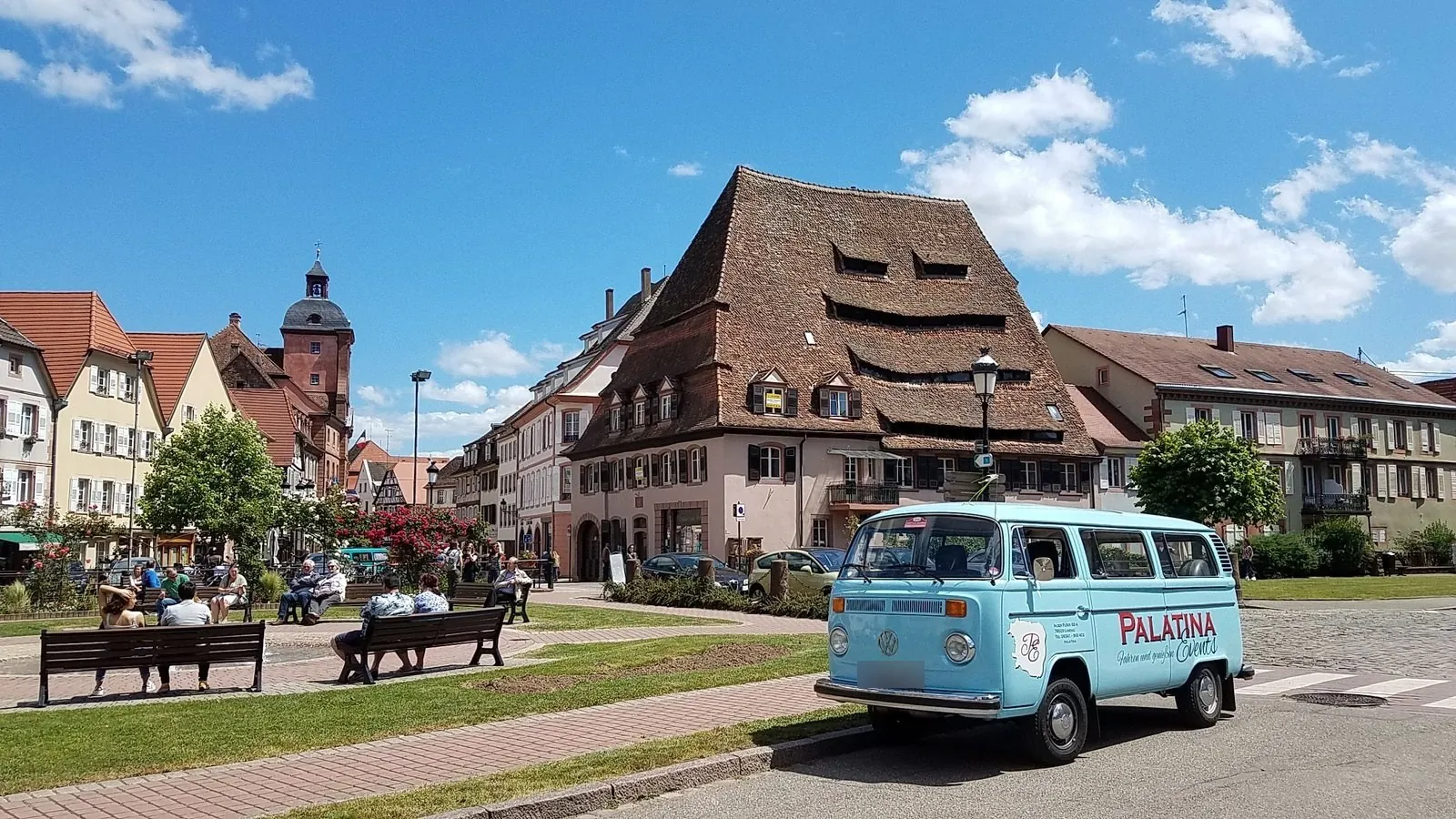 Hellblauer VW Bulli mit Palatina-Schriftzug auf einem Platz vor einem Fachwerkhaus mit hohem Dach