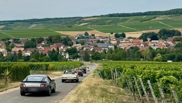 Porsche 928 und weitere Oldtimer fahren hintereinander auf einem schmalen Weg zwischen Reben auf ein Dorf zu