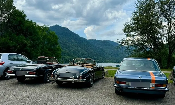 Drei klassische Autos von hinten auf einem Parkplatz an einem See vor bewaldeten Bergen