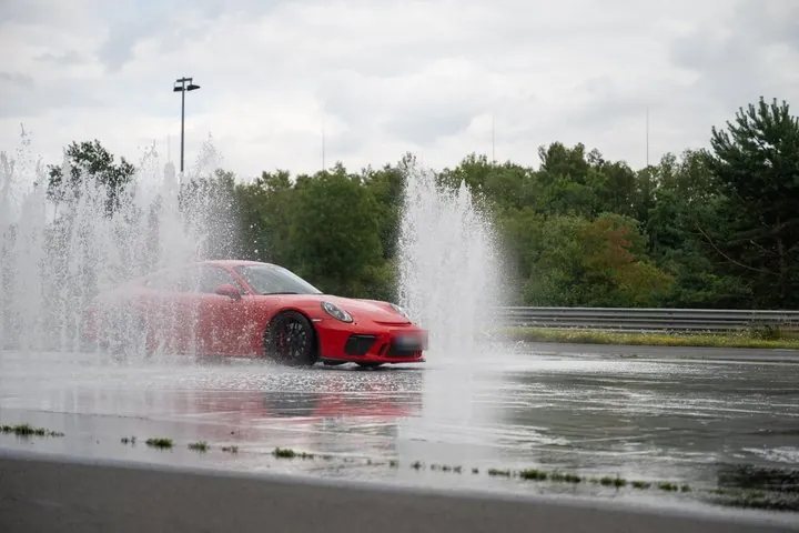 Roter Sportwagen auf einer nassen Trainingsfläche zwischen Wasserfontänen, Kennzeichen unkenntlich