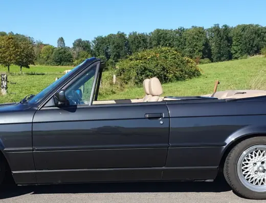 Bmw 320i Baujahr 1990 zu verkaufen – Dunkelblauer BMW 320i Cabriolet mit geöffnetem Verdeck, Leichtmetallfelgen und beige Ledersitzen, geparkt vor grüner Landschaft.