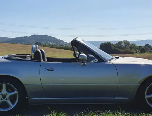Mazda MX5 Baujahr 1990 zu verkaufen – Silbergrauer Mazda MX5 Roadster mit geöffnetem Verdeck in einer ländlichen Landschaft, Seitenansicht von links.
