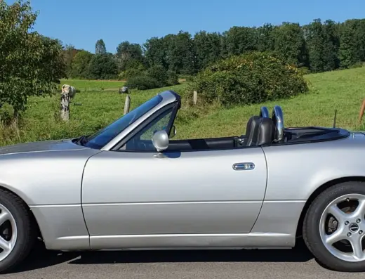 Mazda MX5 Baujahr 1990 zu verkaufen – Silberner Mazda MX5 Roadster mit geöffnetem Verdeck in der Seitenansicht auf einer Landstraße vor grüner Wiese und Baumkulisse.