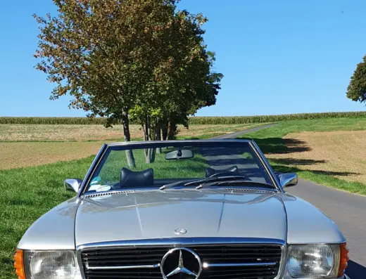 Mercedes Benz 350 SL Baujahr 1971 zu verkaufen – Silberner Mercedes-Benz 350 SL Roadster mit geöffnetem Verdeck auf ländlicher Straße vor Baumgruppe und Feldern.