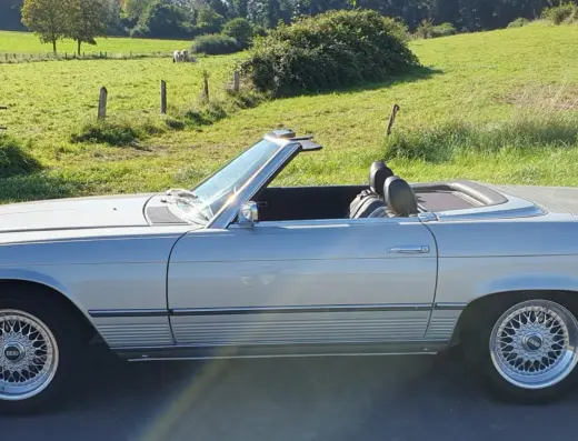 Mercedes Benz 350 SL Baujahr 1971 zu verkaufen – Hellblauer Mercedes-Benz 350 SL Roadster mit geöffnetem Verdeck in Seitenansicht auf einer Landstraße vor grüner Wiese und Feldern.