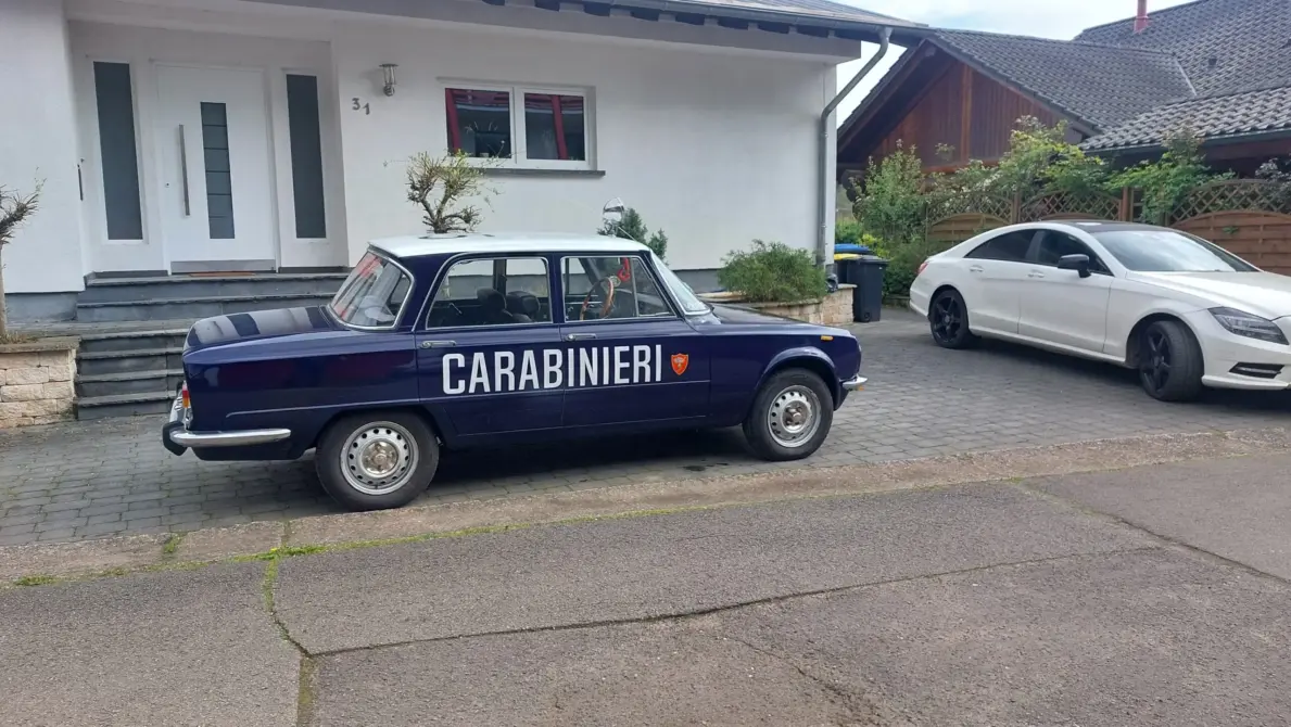 Alfa Romeo Nuova Super 1300 zu verkaufen, Ein dunkelblauer Alfa Romeo Nuova Super 1300 mit "CARABINIERI" Beschriftung steht auf einem gepflasterten Parkplatz vor einem modernen weißen