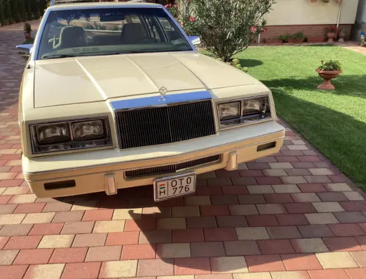 Chrysler Le Baron Baujahr 1982 zu verkaufen – Cremefarbener Chrysler LeBaron von vorne auf gepflasterter Einfahrt, mit senkrecht gegliedertem Kühlergrill und rechteckigen Scheinwerfern