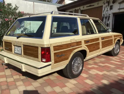 Chrysler Le Baron Baujahr 1982 zu verkaufen – Cremefarbener Chrysler LeBaron Town & Country mit Holzdekor schräg von hinten, vor einer Garage auf Pflaster geparkt, Kennzeichen sichtbar