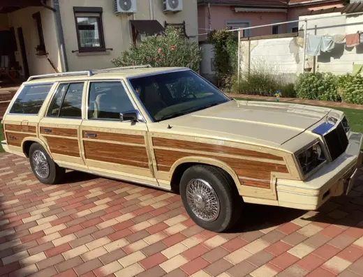 Chrysler Le Baron Baujahr 1982 zu verkaufen – Cremefarbener Chrysler LeBaron Town & Country mit Holzverkleidung, geparkt auf Ziegelpflaster vor einem Wohnhaus.