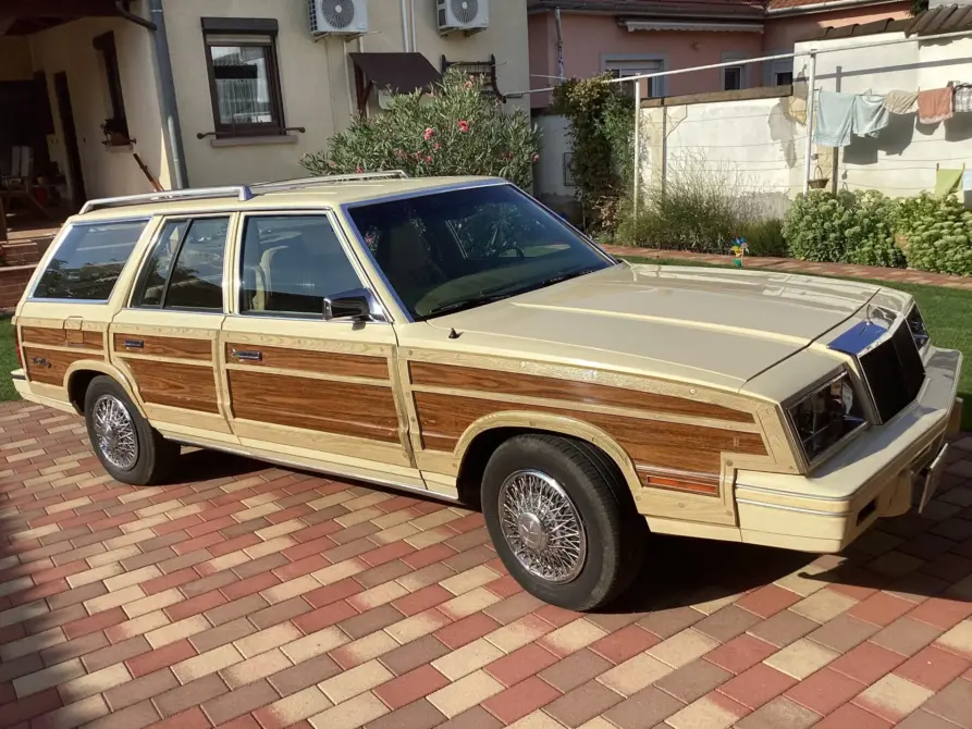 Chrysler Le Baron Baujahr 1982 zu verkaufen, Cremefarbener Chrysler LeBaron Town & Country mit Holzverkleidung, geparkt auf Ziegelpflaster vor einem Wohnhaus.