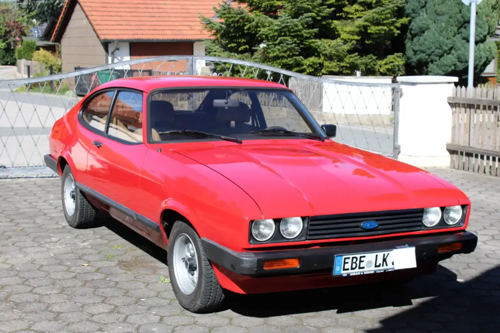 Ford Capri Baujahr 1980 zu verkaufen: Roter Ford Capri Coupé schräg von vorne auf Pflaster, Stoßstange, Scheinwerfer und Räder sichtbar, Gebäude im Hintergrund.