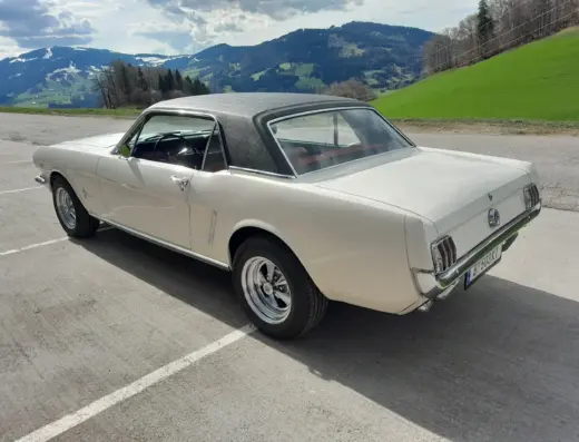 Ford Mustang Baujahr 1965 zu verkaufen – Weißer Ford Mustang Coupé mit schwarzem Vinyldach von schräg hinten auf einem Parkplatz vor Berglandschaft, Fenster offen.