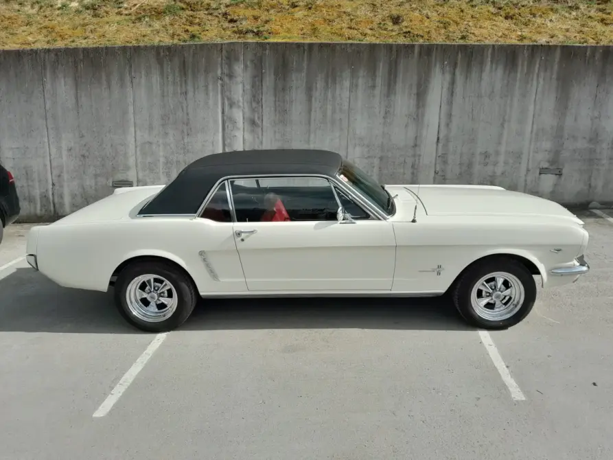 Ford Mustang Baujahr 1965 zu verkaufen, Weißer Ford Mustang Coupé mit schwarzem Vinyldach und rotem Interieur, Seitenansicht auf einem Parkplatz vor einer Betonwand.