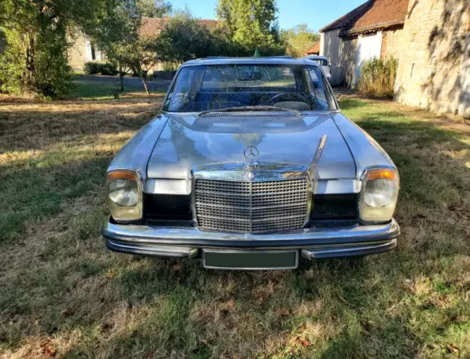 Mercedes Benz 250CE Baujahr 1971 zu verkaufen, Silbergrauer Mercedes-Benz 250CE Coupé von vorne auf einer Wiese vor alten Gebäuden, mit verchromtem Grill und runden Scheinwerfern.