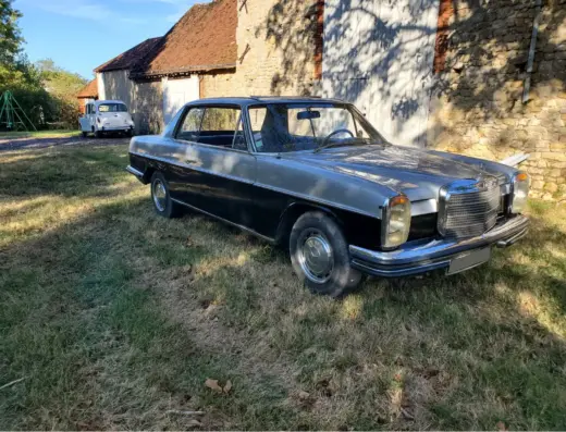 Mercedes Benz 250CE Baujahr 1971 zu verkaufen, Silbernes Mercedes-Benz 250CE Coupé von links auf einer Wiese vor einem Steingebäude, Chromzierleisten und runde Scheinwerfer sichtbar.
