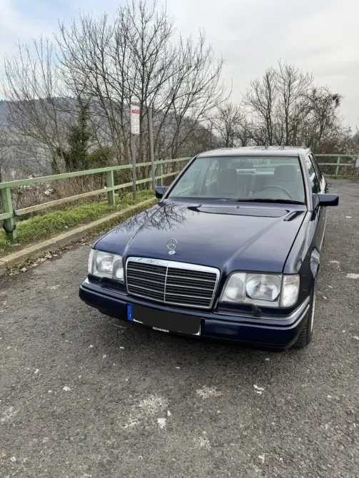 Mercedes Benz E280 Baujahr 1995 zu verkaufen, Dunkelblaue Mercedes-Benz E280 Limousine von vorne auf einem Parkplatz vor grünem Metallgeländer und kahlen Bäumen.