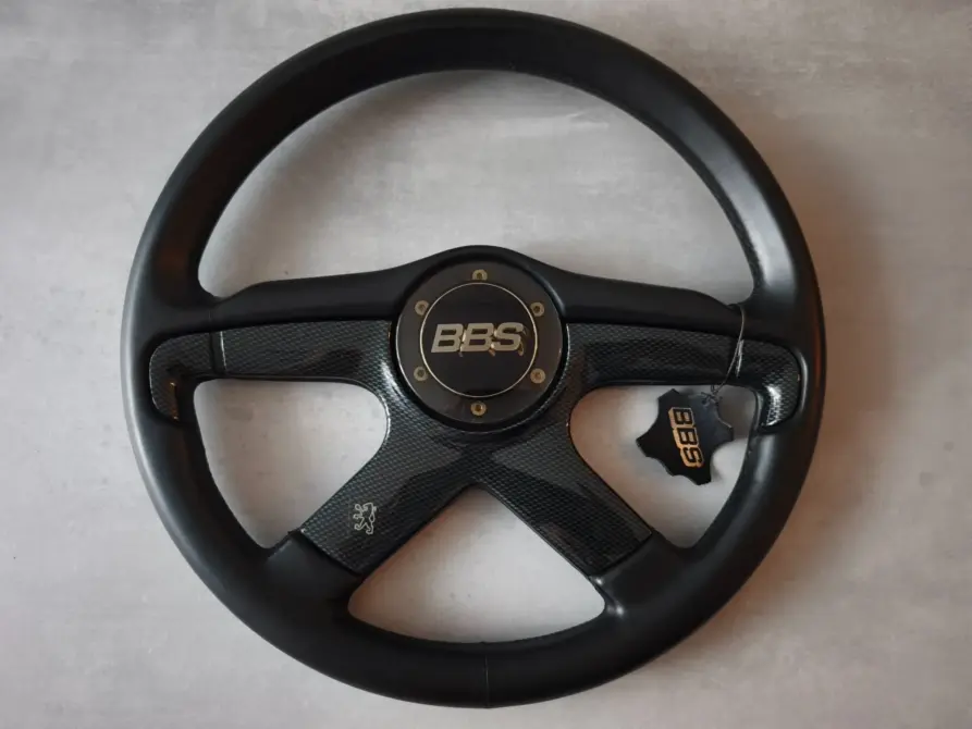 BBS Italvolanti Sportlenkrad, Typ zu verkaufen: Schwarzes Vierspeichen-Sportlenkrad mit BBS Emblem in der Mitte und Carbon-Optik an den Speichen, liegt auf hellgrauem Untergrund
