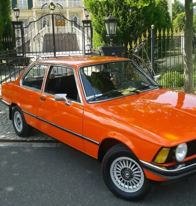 BMW 316 Baujahr 1976 zu verkaufen: Orange BMW 316 Limousine schräg von vorne rechts auf Asphalt, Rad, Scheinwerfer und Seitenspiegel sichtbar