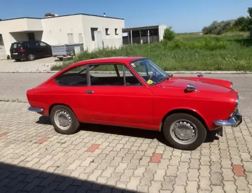 Fiat 850 Sport Coupé Baujahr 1971 zu verkaufen: Roter Fiat 850 Sport Coupé steht auf Pflastersteinen, Gebäude und Wiese im Hintergrund sichtbar.