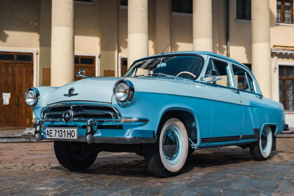 GAZ-21 Wolga Baujahr 1958 zu verkaufen: Blaue zweifarbige Limousine schräg von vorn links, Kühlergrill mit Stern und Hirschfigur auf der Motorhaube, vor einem Säulengebäude