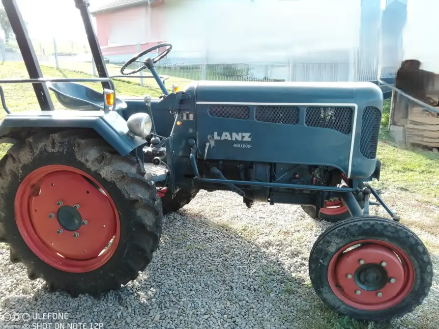 Lanz D1666 Baujahr 1956 zu verkaufen: Blauer Lanz Bulldog Traktor mit roten Felgen von der rechten Seite auf Kies, Lenkrad, Sitz und Scheinwerfer sichtbar, Wiese dahinter.