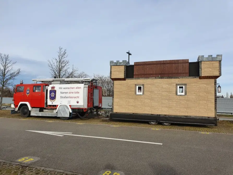 Magirus Deutz LF 16 TS Baujahr 1981 zu verkaufen: Rotes Feuerwehrfahrzeug mit Banner an der Seite, Anhänger mit Burgaufbau daneben, auf Asphalt und Pflaster im Freien