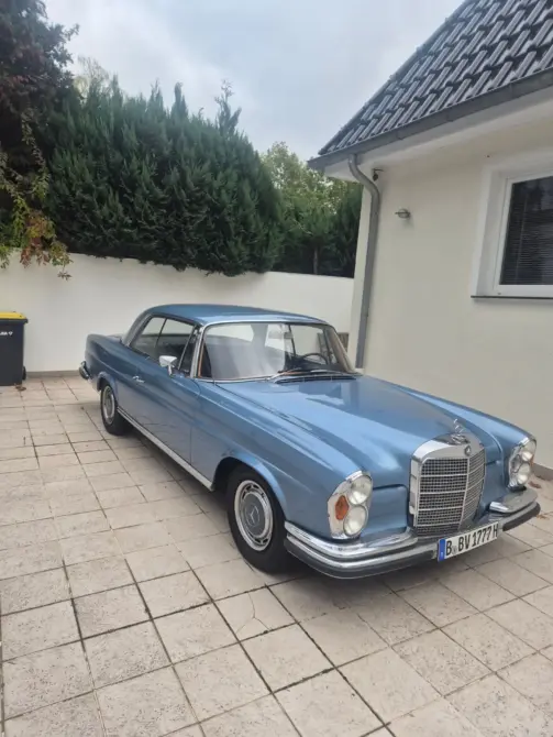 Mercedes-Benz 280 SE Baujahr 1969 zu verkaufen: Blaues Coupé schräg von vorne rechts auf Pflaster, Kühlergrill, Scheinwerfer und Stoßstange sichtbar, daneben Hauswand und Hecke