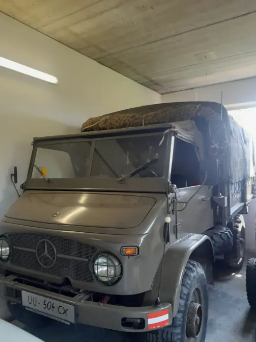 Mercedes-Benz Unimog 404 S Baujahr 1964 zu verkaufen: Olivgrüner Unimog von schräg vorne in einer Garage, Mercedes-Stern im Kühlergrill, Plane über der Ladefläche