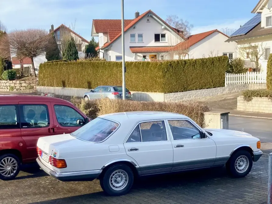 Mercedes-Benz W126.032 S380 zu verkaufen: Weiße Limousine seitlich von links auf Pflaster, dahinter ein weiteres Auto, Hecke und Wohnhäuser