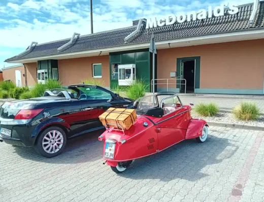 Messerschmitt KR 200 Cabrio zu verkaufen, roter Kabinenroller schräg von hinten mit H-Kennzeichen und Koffer auf dem Gepäckträger, daneben ein modernes Cabrio
