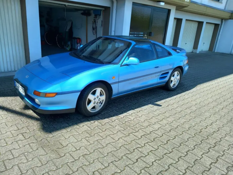 Toyota MR2 Baujahr 1993 zu verkaufen: Blauer Toyota MR2 Coupé schräg von vorne links auf Pflaster vor Garagen, Felgen und Seitenspiegel sichtbar