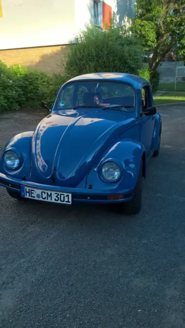 Volkswagen Mexiko Käfer 1600i Baujahr 1996 zu verkaufen: Blauer VW Käfer schräg von vorne links auf Asphalt, Scheinwerfer, Haube und Kennzeichen sichtbar, dahinter Büsche und Gebäude