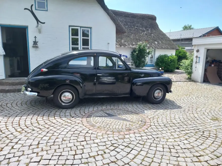 Volvo PV444 Baujahr 1954 zu verkaufen: Schwarze Volvo PV444 Limousine von der Seite auf Kopfsteinpflaster vor weißem Reetdachhaus, Front zeigt nach rechts